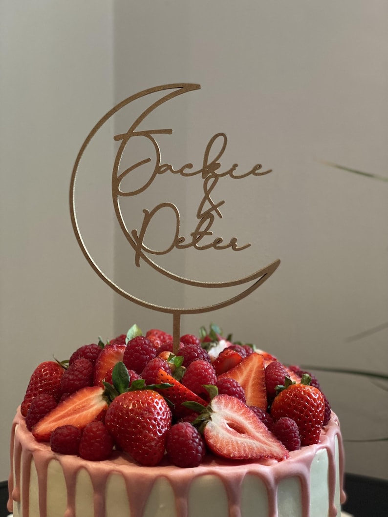 Op de afbeelding: Een witte cake met roze glazuur en verse aardbeien en frambozen. Een houten caketopper in de vorm van een halve maan met de namen "Jackie & Peter" erop.