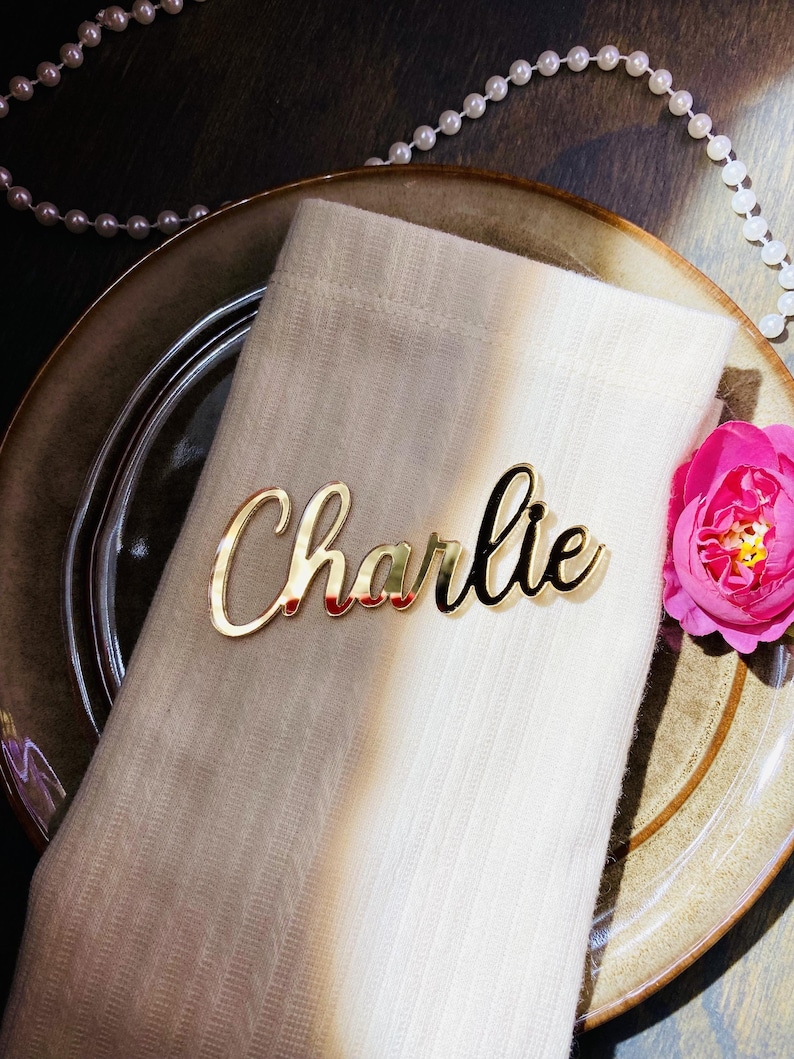 May include: A gold-colored nameplate that reads "Charlie" in cursive script rests on a folded white napkin. The napkin is placed on a decorative plate, with a pink flower and a string of pearls in the background. The nameplate is a decorative element.