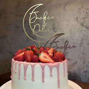 Op de afbeelding: Een witte cake met roze glazuur en een gouden maan-topper met de namen "Jackie & Peter" erop. De cake is versierd met verse aardbeien en frambozen.