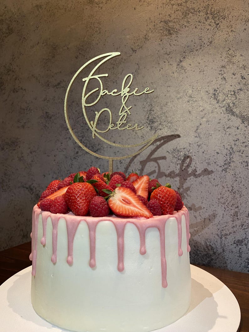Op de afbeelding: Een witte cake met roze glazuur en een gouden maan-topper met de namen "Jackie & Peter" erop. De cake is versierd met verse aardbeien en frambozen.