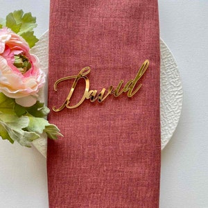 Puede incluir: Tarjeta de lugar de acr&iacute;lico dorado con el nombre "David" escrito en cursiva. La tarjeta de lugar est&aacute; sobre una servilleta de lino roja.