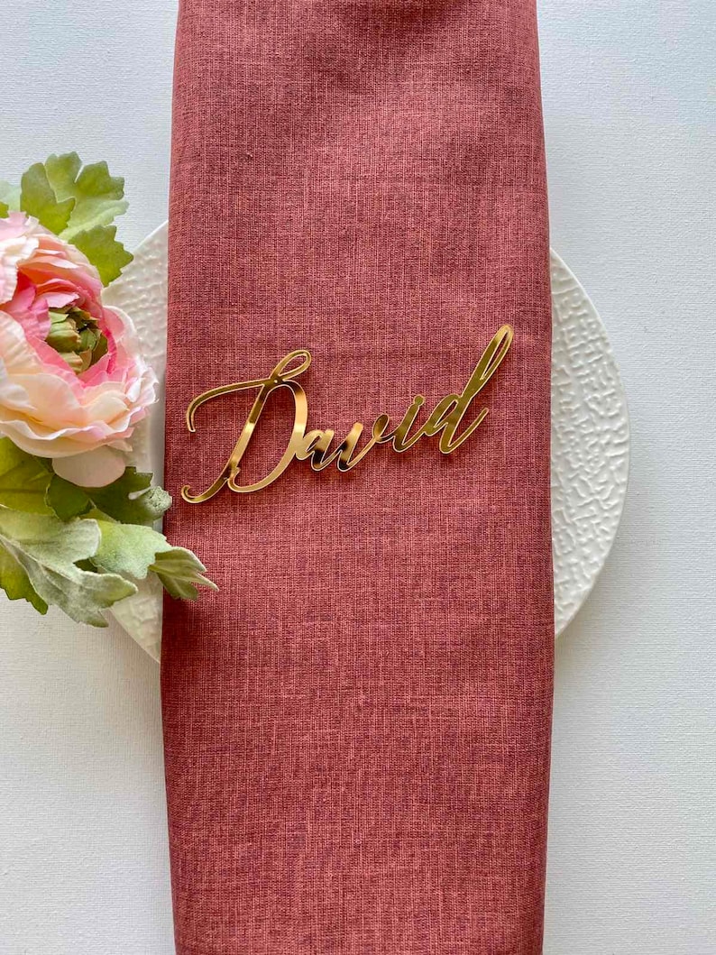 Puede incluir: Tarjeta de lugar de acr&iacute;lico dorado con el nombre "David" escrito en cursiva. La tarjeta de lugar est&aacute; sobre una servilleta de lino roja.