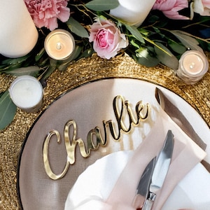 Puede incluir: Una mesa decorada con un identificador dorado con el nombre "Charlie" en cursiva. El plato está adornado con una servilleta blanca, cubiertos y un lazo rosa. La mesa está decorada con flores rosas, vegetación y velas encendidas.