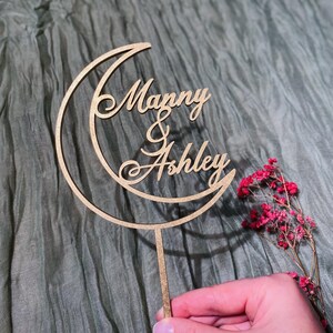 Op de afbeelding: Een houten taarttopper in de vorm van een halve maan met de namen "Manny & Ashley" in cursief.