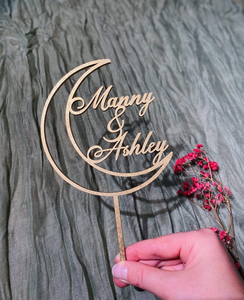 Op de afbeelding: Een houten taarttopper in de vorm van een halve maan met de namen "Manny & Ashley" in cursief.