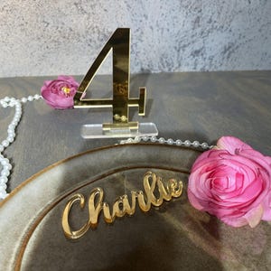 May include: A gold acrylic table number 4 stands on a clear base. A gold acrylic nameplate that says "Charlie" rests on a brown plate. Pink roses and a string of pearls are also present.