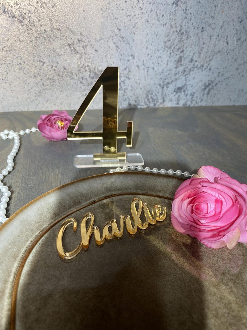 May include: A gold acrylic table number 4 stands on a clear base. A gold acrylic nameplate that says "Charlie" rests on a brown plate. Pink roses and a string of pearls are also present.