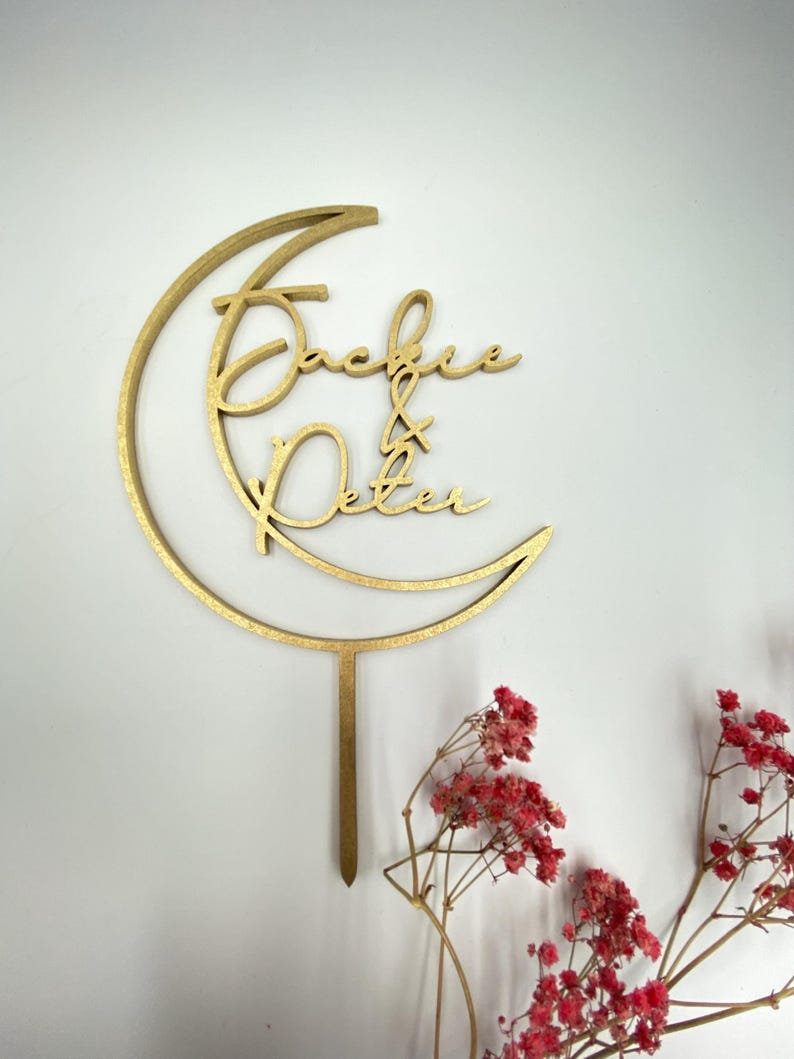 Op de afbeelding: Gouden houten taarttopper in de vorm van een halve maan met de namen "Jackie & Peter" in cursief schrift.