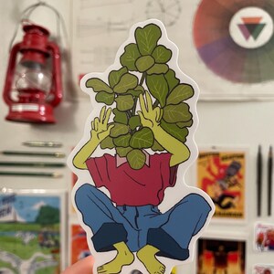 Peut inclure: Un autocollant représentant une personne assise avec la tête recouverte d'une grande plante verte. La personne porte une chemise rouge et un jean bleu. L'autocollant est sur un fond blanc.