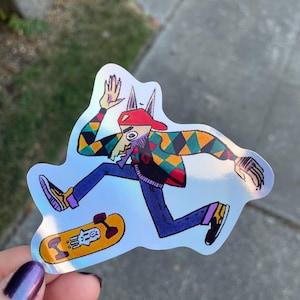 Puede incluir: Una pegatina holográfica con un personaje de dibujos animados con cara de lobo, gorra roja y chaqueta estampada de colores. El personaje anda en monopatín sobre una patineta amarilla con un diseño blanco.