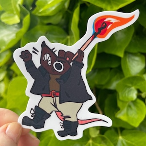 Puede incluir: Una pegatina con un personaje de rata de dibujos animados con una chaqueta negra, pantalones color beige y botas negras, sosteniendo un fósforo encendido. La rata tiene una cara marrón con un ojo grande, sobre un fondo verde.