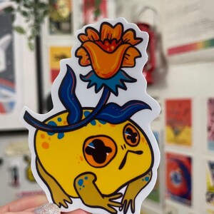Kleiner Frosch mit einem Blumenhut-Vinylaufkleber, wasserfest