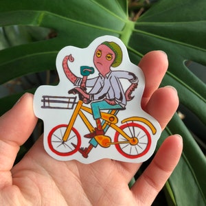 May include: A sticker of an octopus riding a yellow bicycle with red wheels. The octopus is wearing a blue shirt, blue jeans, and a green beanie. The octopus has yellow eyes and a pink body.
