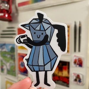 May include: A sticker featuring a cartoon-style blue and gray coffee maker holding a small white coffee cup. The coffee maker has a smiling face and black handle. The sticker is held up against a white surface with other stickers and a red lantern in the background.
