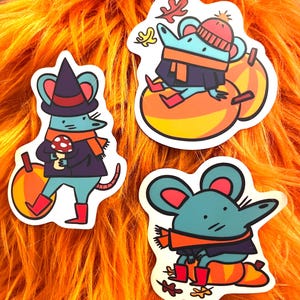 Puede incluir: Tres pegatinas caprichosas con ratones de dibujos animados con atuendos otoñales. Un ratón lleva un sombrero de bruja, otro un gorro de punto, todos con bufandas y botas. Las pegatinas están sobre un fondo naranja vibrante.