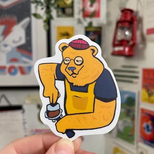 May include: A cartoon sticker of a bear wearing a red beanie and a yellow apron, holding a coffee machine. The bear has a serious expression and is making coffee.