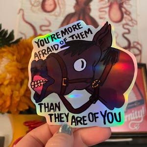 Puede incluir: Pegatina holográfica con la cabeza de un caballo de dibujos animados con brida. El texto de la pegatina dice: "Les tienes más miedo que ellos a ti". El caballo es marrón con la boca rosa.