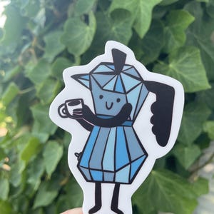 May include: A sticker depicting a cartoon blue moka pot with a smiling face, holding a coffee cup. The moka pot is wearing a dress. The sticker is white, set against a green leafy background.