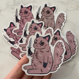 Puede incluir: Una colección de pegatinas con un gato de dibujos animados con una expresión severa, sosteniendo un cuchillo. El gato es de color malva con acentos negros, y el cuchillo tiene una hoja plateada y un mango negro. Las pegatinas están sobre un fondo blanco.