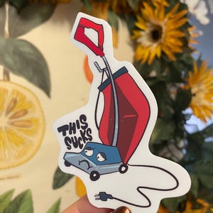 May include: A sticker featuring a cartoon vacuum cleaner with a red bag and blue body. The text "THIS SUCKS" is above the vacuum. The sticker is held by a person with a background of sunflowers and a botanical print.