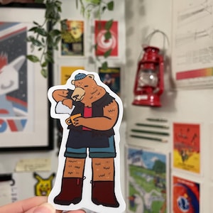 May include: A brown bear wearing a blue hat, blue shorts, and brown boots is holding a cup of coffee. The bear is looking down at the cup. The sticker is on a white background.