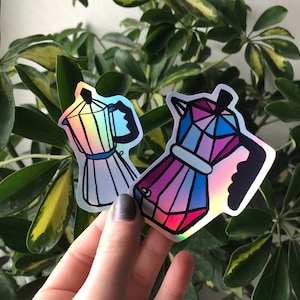 Könnte beinhalten: Zwei regenbogenfarbene, holografische Aufkleber von Kaffeekannen. Die Kaffeekannen sind in einem Regenbogen-Farbschema mit einem blauen, rosa und violetten Design gehalten.