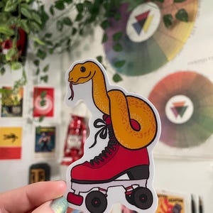 Puede incluir: Una pegatina con una serpiente de dibujos animados naranja que lleva un patín rojo. La serpiente tiene un contorno blanco y una lengua bífida. El patín es rojo con detalles negros y ruedas negras. La pegatina la sostiene una persona.