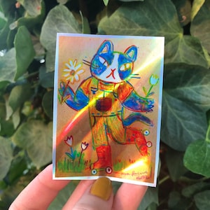 May include: A holographic sticker featuring a cartoon cat wearing overalls and roller skates. The cat is holding a daisy and there are other flowers in the background. The sticker has a rainbow iridescent finish.