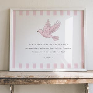 May include: A framed print featuring a pink dove illustration with text from Matthew 6:26. The artwork has a white frame with a pink striped border. The print is displayed on a rustic wooden shelf against a neutral wall.