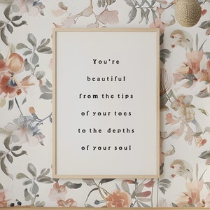 May include: A white crib with a white mattress and a pink pillow. The crib is in a room with a floral wallpaper and a framed print that reads "You're beautiful from the tips of your toes to the depths of your soul."