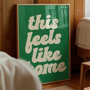 May include: A framed art print with a green background and the words "this feels like home" in a white, retro font. The artwork is displayed in a room with wooden paneling and a bed.