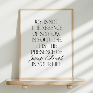 May include: A framed print with a light wood frame displays the text "JOY IS NOT THE ABSENCE OF SORROW IN YOUR LIFE. IT IS THE PRESENCE OF Jesus Christ IN YOUR LIFE." The print rests on a light wood shelf against a white wall.