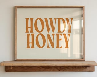 May include: A framed print with the words "HOWDY HONEY" in orange lettering. The print is displayed on a wooden shelf against a white wall. The frame is a light brown color.