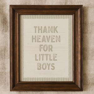 Puede incluir: Un marco de madera marr&oacute;n oscuro que contiene una impresi&oacute;n con el texto "THANK HEAVEN FOR LITTLE BOYS" en gris. El fondo es beige claro con un patr&oacute;n sutil. Es un objeto decorativo.