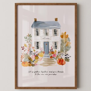 May include: A framed watercolor print of a white house with a blue door and shutters, surrounded by pumpkins and fall foliage. The word "welcome" is above the door. The bottom of the print has the text "Let us gather together and give thanks to the one who provides."