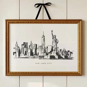 May include: A framed black and white print of the New York City skyline, including the Statue of Liberty, with the text "NEW YORK CITY." The gold frame is suspended by a black ribbon tied in a bow.