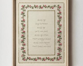 May include: Framed embroidery with the text "Bless My Cozy Kitchen" surrounded by a floral border of pink roses and green leaves. The frame is a light beige color, and the background is a cream color.
