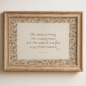 May include: A rectangular wooden frame containing a cream-colored paper with the text: The grass withers, the flower fades, but the word of our God will stand forever. Isaiah 40:8. The paper is bordered by a small, multi-colored floral pattern.