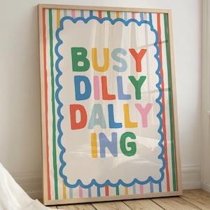 May include: A framed art print with the words "BUSY DILLY DALLY ING" in colorful block letters. The print has a cream background with a blue scalloped border and colorful vertical stripes. The frame is made of light-colored wood.