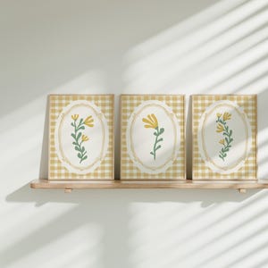 May include: Three framed prints featuring a yellow and green floral design on a white background. The frames are a light wood colour and are displayed on a wooden shelf.