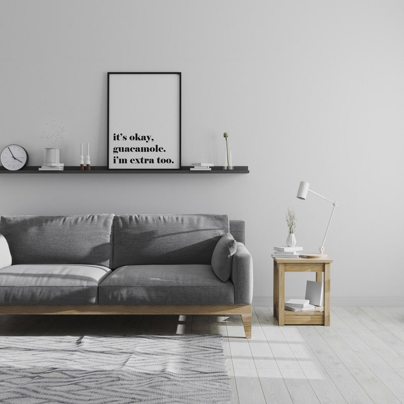 Puede incluir: Un sof&aacute; gris con un marco de madera se encuentra frente a una pared blanca. Una repisa negra con una impresi&oacute;n enmarcada que dice "it's okay, guacamole, I'm extra too." est&aacute; montada sobre el sof&aacute;. Una peque&ntilde;a mesa auxiliar de madera con una l&aacute;mpara blanca y un jarr&oacute;n de flores se encuentra a la derecha del sof&aacute;.