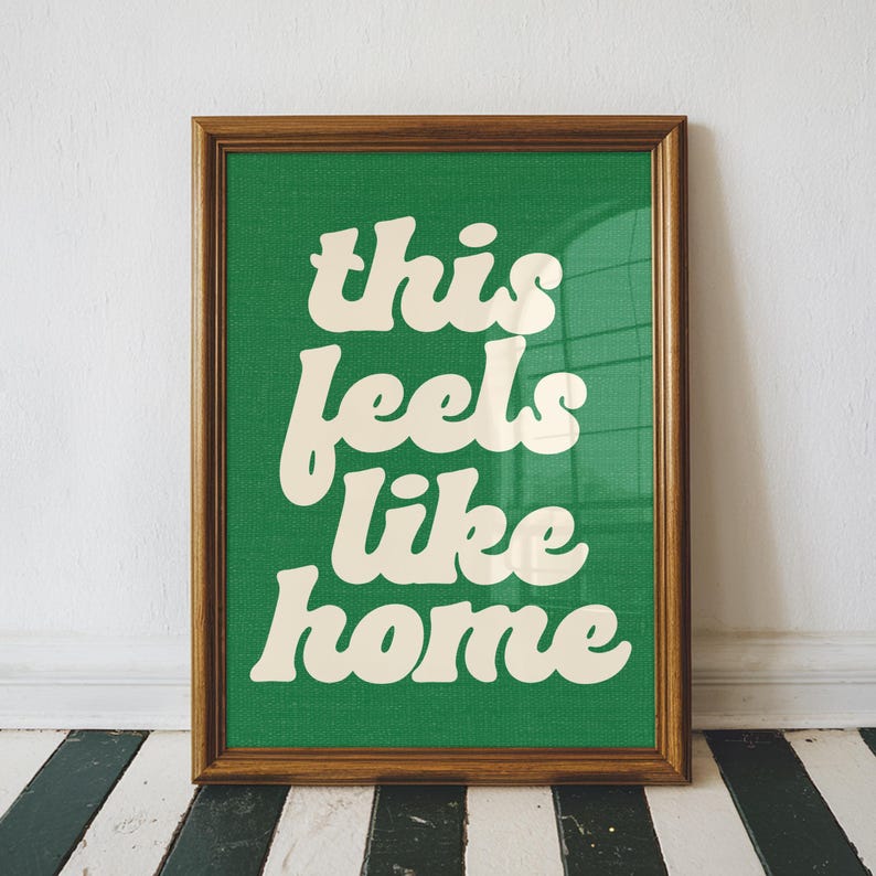 May include: A framed art print with the phrase "this feels like home" in a white, retro font. The text is set against a green textured background, encased in a brown wooden frame. The artwork is displayed against a white wall and a black and white striped floor.