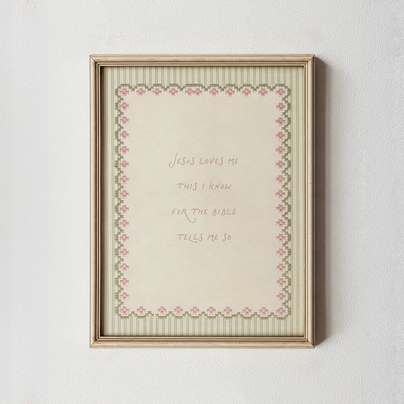May include: A framed cross-stitch sampler featuring the text "Jesus loves me this I know for the Bible tells me so" in brown lettering. The design includes a decorative border of small pink flowers and green leaves on a cream background.