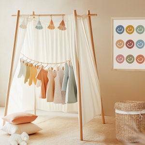 May include: A wooden clothing rack with a white curtain draped over it. The rack is decorated with colorful tassels and has a variety of children's clothing hanging from it. A framed print with colorful smiley faces hangs on the wall behind the rack. A wicker basket sits on the floor in front of the rack.