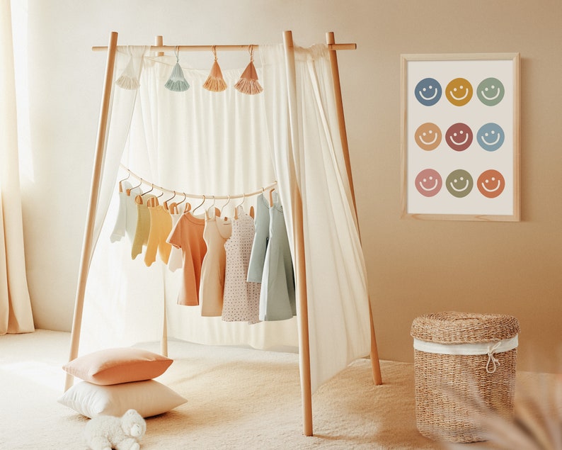 May include: A wooden clothing rack with a white curtain draped over it. The rack is decorated with colorful tassels and has a variety of children's clothing hanging from it. A framed print with colorful smiley faces hangs on the wall behind the rack. A wicker basket sits on the floor in front of the rack.