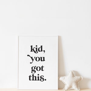 May include: A white framed print with black text that reads "kid, you got this." The print is on a light brown surface with a white star-shaped pillow.