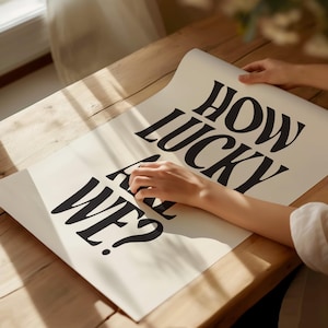 May include: A white poster with black text that reads "How lucky are we?" The poster is being held by a person's hand.