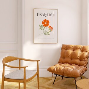 K&ouml;nnte beinhalten: Ein gerahmter Kunstdruck mit dem Text "PSALM 16:11" und einer floralen Illustration in Orange und Gr&uuml;n. Der Druck h&auml;ngt an einer wei&szlig;en Wand &uuml;ber einem braunen Ledersessel und einem Holzstuhl mit einem grauen Kissen. Vor dem Stuhl steht ein kleiner Holztisch.