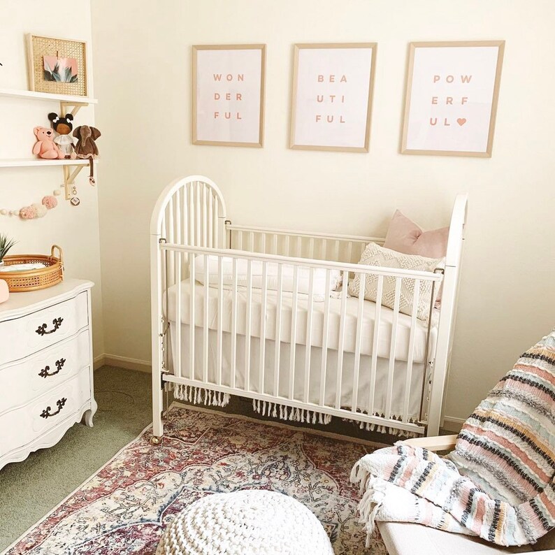 May include: A white crib with a white mattress and a pink and white blanket. There are three framed prints on the wall behind the crib, each with the word "ful" in pink letters. The prints read "Wonderful", "Beautiful", and "Powerful".