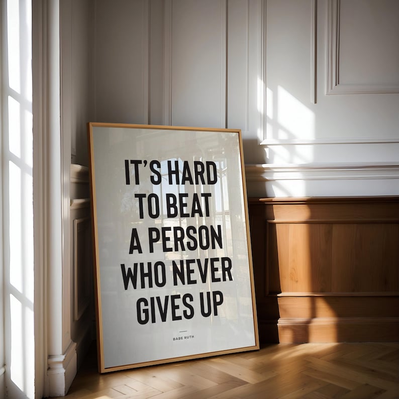 Puede incluir: Un cuadro enmarcado con la cita "IT'S HARD TO BEAT A PERSON WHO NEVER GIVES UP" en letras negras. El cuadro tiene un marco de madera clara y est&aacute; apoyado contra una pared blanca y un panel de madera. La cita se atribuye a Babe Ruth.